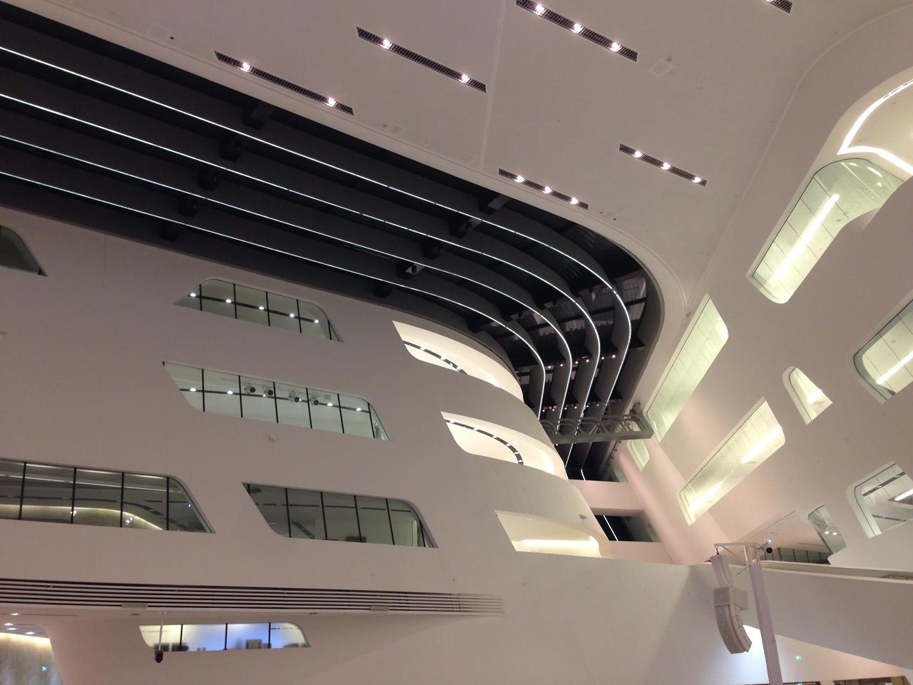Moderne Innenarchitektur eines großen, hellen öffentlichen Raumes mit geschwungenen weißen Wänden, gestaffelter Beleuchtung und horizontalen Fenstern.