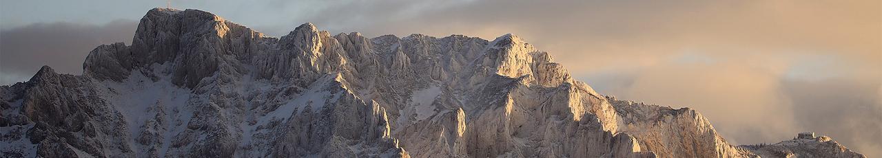 Snow-covered mountain range at sunrise or sunset, with dramatic lighting and fog in the valleys.